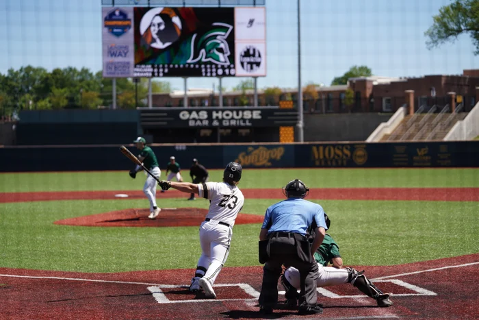 A baseball batter swinging as the catcher and umpire watch from behind home plate